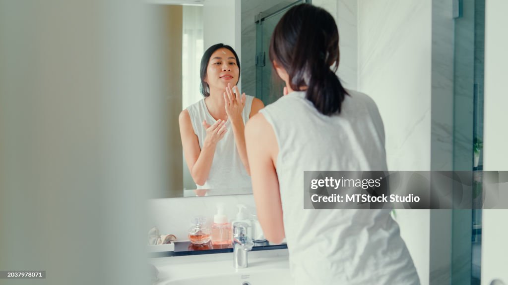 Young Asian woman having fun apply facial cream look in mirror in lavatory at home late morning. Routine lifestyle in house.