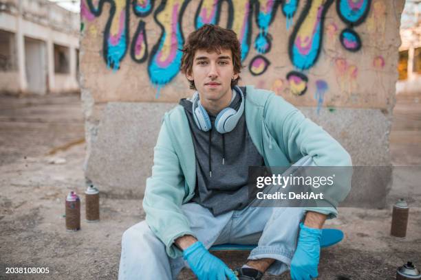 a young man looks directly into the camera while sitting in front of a graffiti he has drawn - multi coloured glove stock pictures, royalty-free photos & images