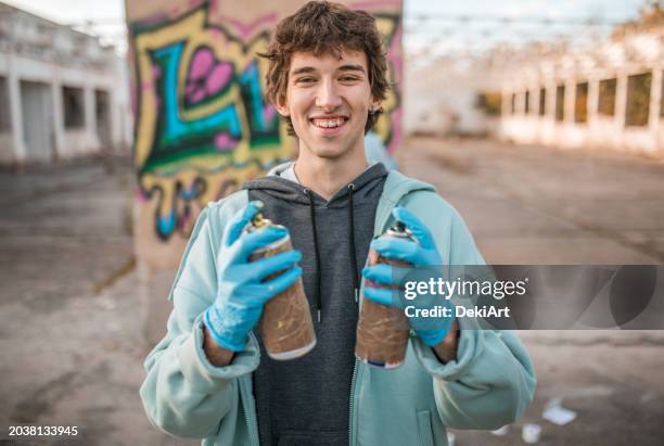 porträt eines lächelnden jungen künstlers, der sprays in den händen hält und es genießt, graffiti zu zeichnen - straßenmaler stock-fotos und bilder