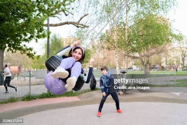 girl sitting on a wheel hanging from a tree in a public park laughing and looking at the camera with her brother behind her, front view - boy girl stock pictures, royalty-free photos & images