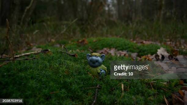 a green troll toy with pink hair and a blue shirt is lying on a bed of moss in a forest. the toy looks happy and relaxed among the natural surroundings - troll fictional character stock pictures, royalty-free photos & images
