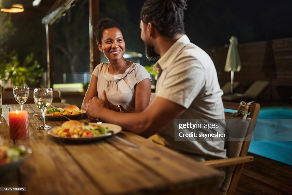 Happy black couple talking during a late meal on a patio by the pool.