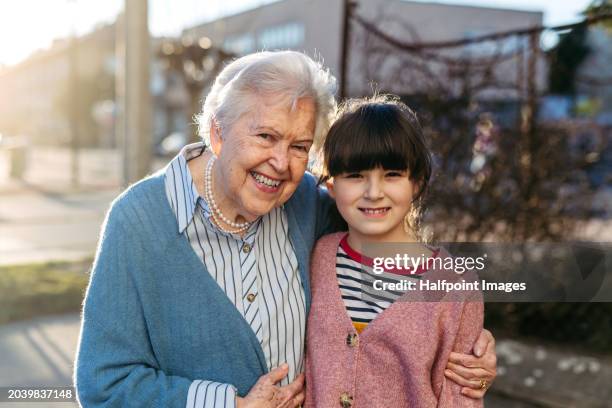 portrait of granddaughter and senior grandmother standing outdoors. girl embracing elderly woman, looking at camera, smiling. - world kindness day stock-fotos und bilder