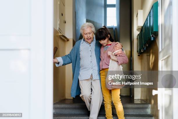 granddaughter helping senior grandmother walk up the stairs, going grocery shopping. girl holding hand of her elderly woman, providing stability while walking. - world kindness day stock-fotos und bilder