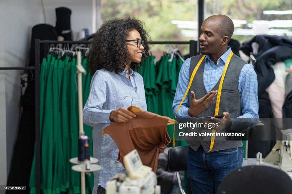 Focused young african-american female tailor in a design studio showing a textile product to a male designer, having discussion among colleagues, fashion ideas exchange and teamwork