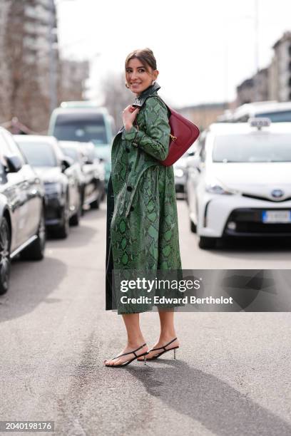 Guest wears golden earrings, a black long dress, a green leather snake print long trench long coat, sandals, outside Ferragamo, during the Milan...