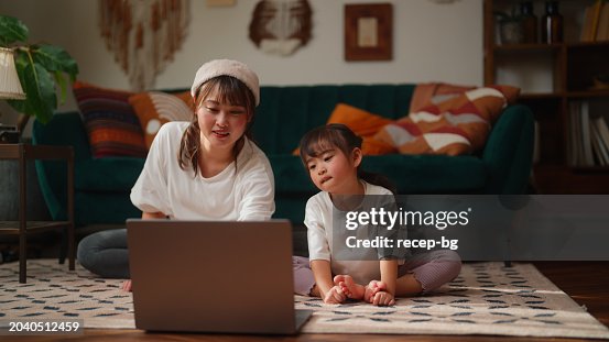 Mother using her laptop to watch online yoga classes and practice with her small daughter in the living room at home Mother using her laptop to watch online yoga classes and practice with her small daughter in the living room at home