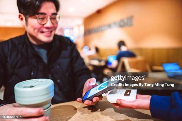 cheerful asian man paying contactless with mobile phone app on wireless terminal in cafe - logic vs emotion stock pictures, royalty-free photos & images