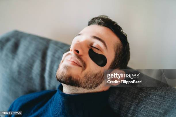 a young adult man is wearing an under eye patch for his daily beauty routine at home - man eye cream imagens e fotografias de stock
