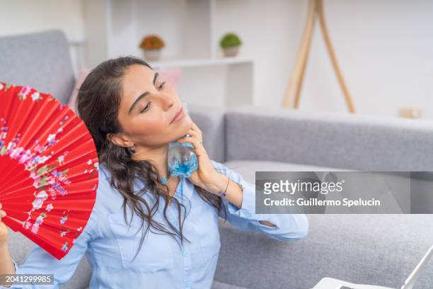 young woman with heat putting a bottle of ice water on her neck and using a fan - ac weary stock pictures, royalty-free photos & images