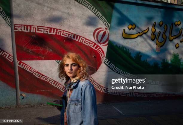 Young woman, not wearing a mandatory headscarf, is walking past a mural featuring an Iranian flag on Enghelab Avenue in downtown Tehran, Iran, on...