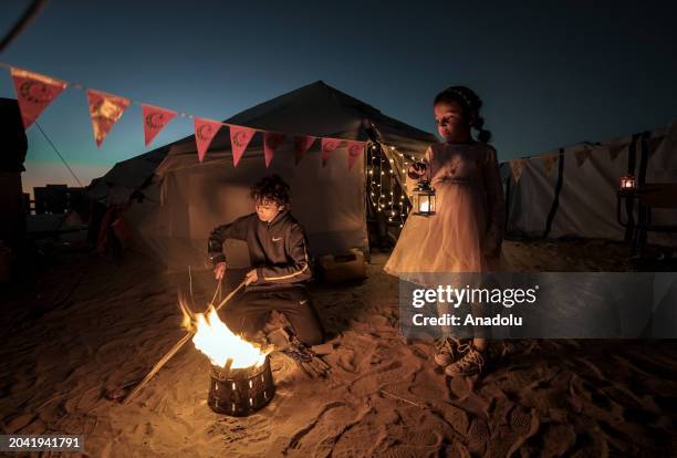 Palestinian boy lights a fire next to a girl holding a Ramadan lantern in front of a tent decorated illuminate lights by Palestinians taking refuge...