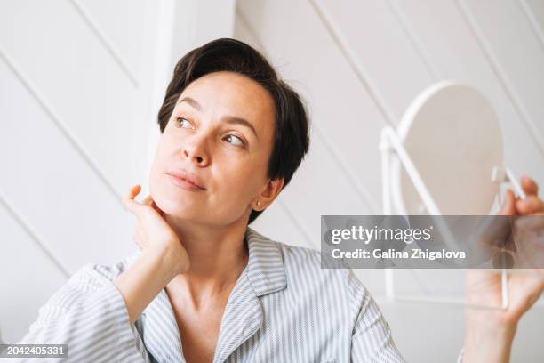 portrait of young adult brunette woman waking up and looking to mirror in bedroom at home, morning skincare routine - brunette woman bedroom stock-fotos und bilder