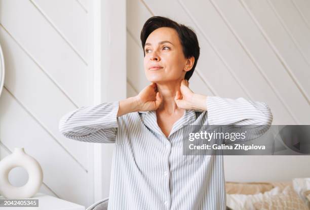 portrait of young adult brunette woman doing facial massage with hands in bedroom at home, morning skincare routine - hals stock-fotos und bilder