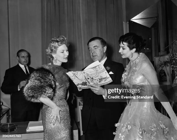 Designer Norman Hartnell looks at his autobiographical book 'Silver and Gold' with fashion models Lana and Cassandra, November 17th 1955.