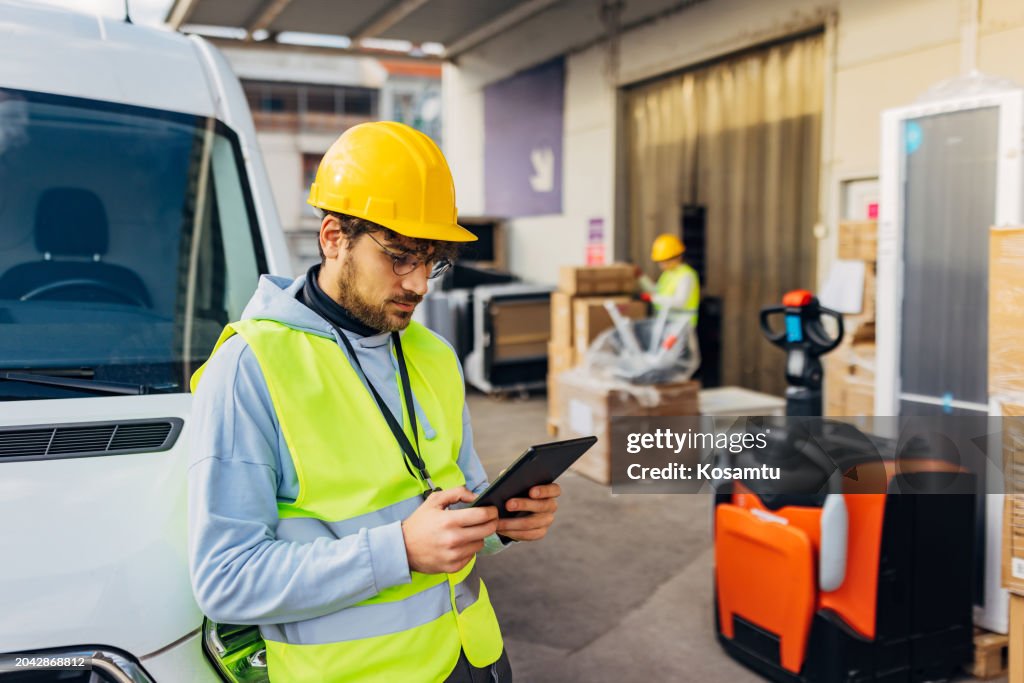 A male warehouse worker stands in front of a wholesale warehouse and uses a digital tablet to check packages for dispatch