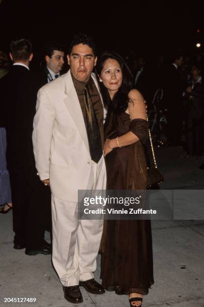 American actor Nicholas Turturro, wearing a white suit over a brown shirt and tie, and his wife, Lissa Espinosa, who wears a brown evening gown and...