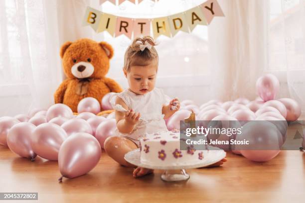 the adorable baby girl is sitting on the floor and eating  her delicious first birthday cake with a spoon. cake smash. - smash cake stockfoto's en -beelden