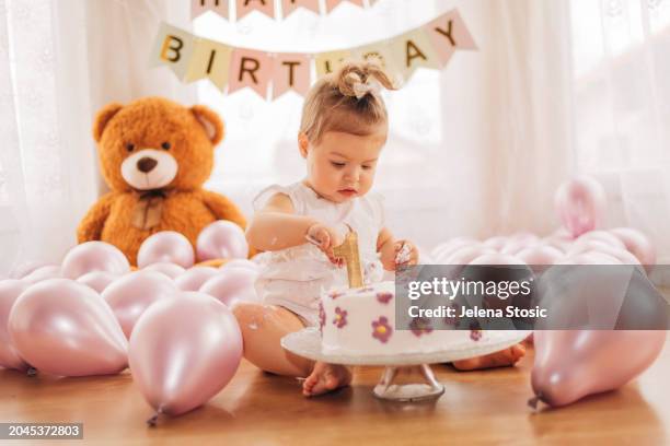 the adorable baby girl is sitting on the floor and eating  her delicious first birthday cake with a spoon. cake smash. - smash cake stockfoto's en -beelden