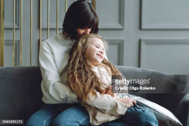 mother and daughter on couch: mother hugs and kisses daughter on her lap - auf dem schoß stock-fotos und bilder