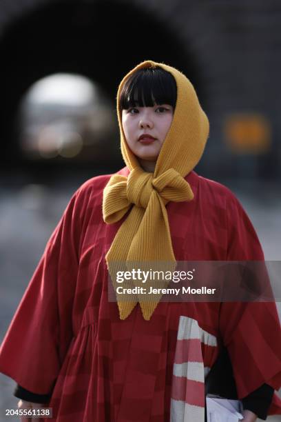 Guest wears a yellow head scarf / piece with a bow detail, red / white striped oversized kimono inspired long coat, outside Anrealage, during the...