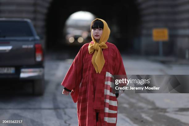 Guest wears a yellow head scarf / piece with a bow detail, red / white striped oversized kimono inspired long coat, outside Anrealage, during the...