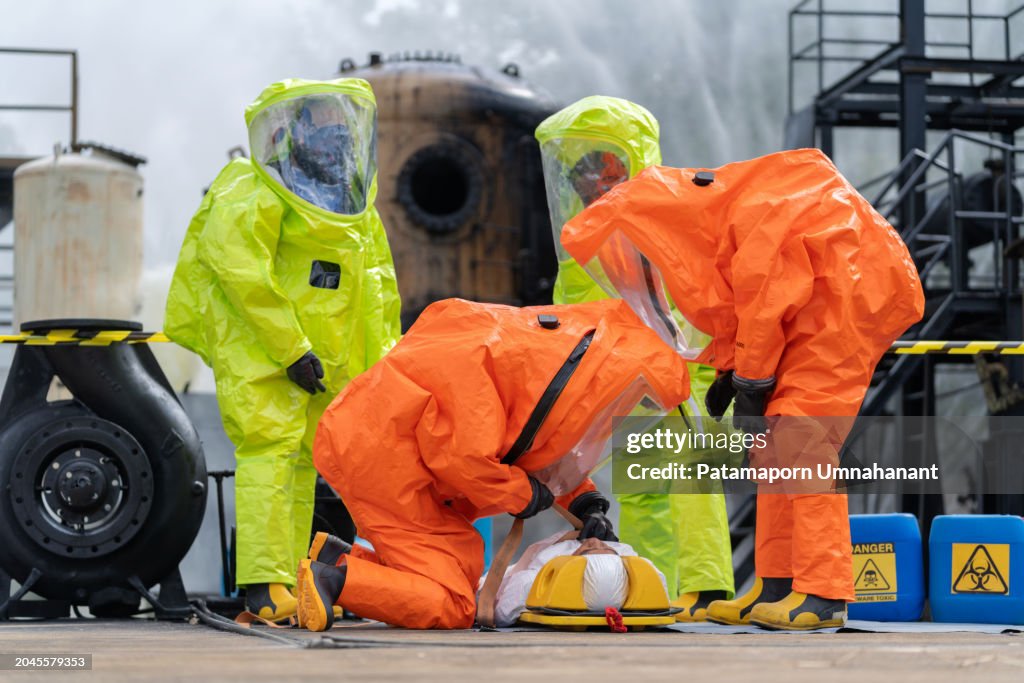 Team of worker, clad in protective gear including a gas mask and hazmat suit, rushes a patient on a field bed away from the hazardous area surrounding a chemical spill from a factory. Teamwork involved in the evacuation are palpable.