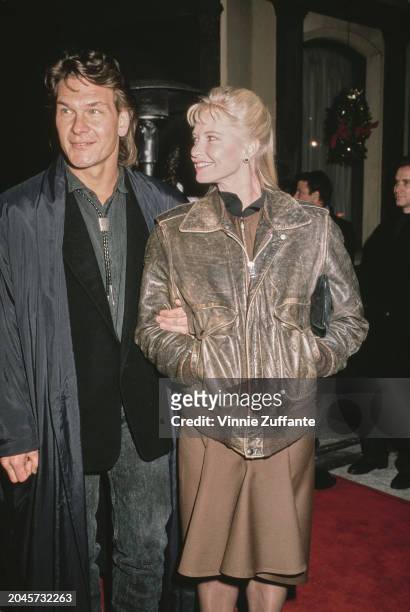 American actor, singer and dancer Patrick Swayze, wearing a grey raincoat over a black jacket with a grey shirt and bolo tie, and his wife, American...