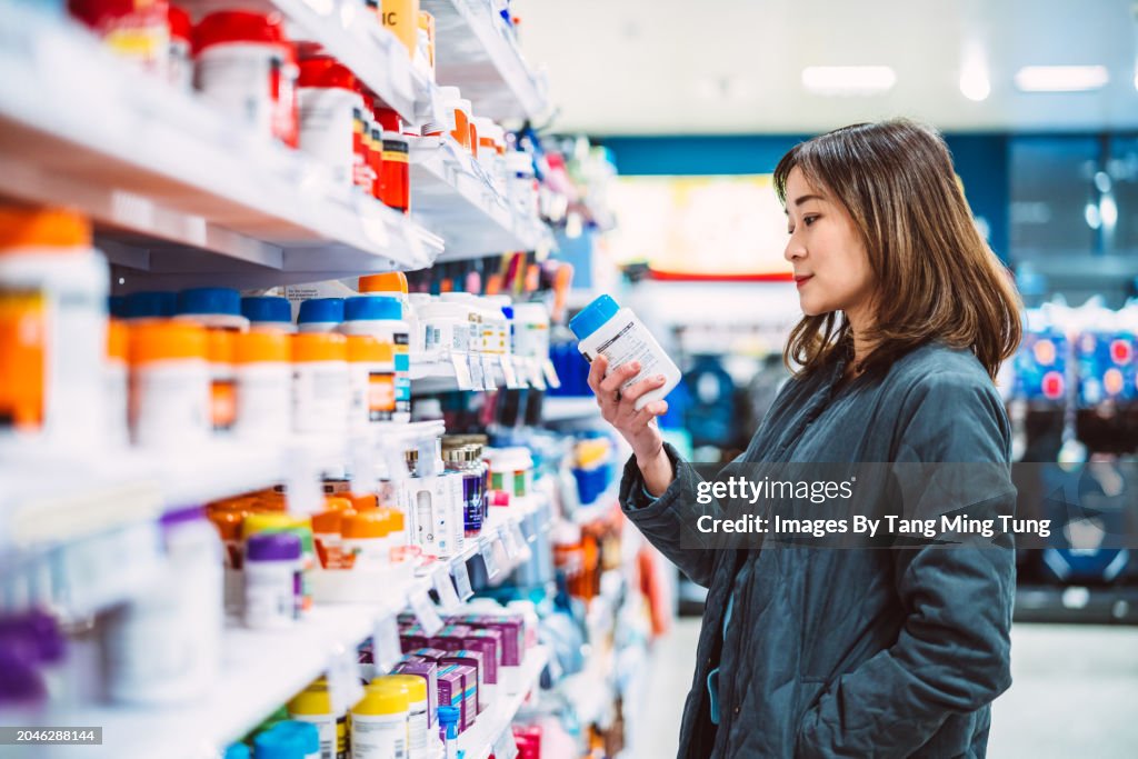 Beautiful Asian woman shopping for multi vitamin & health supplements on shelf in supermarket. Heathy lifestyle concept.