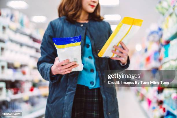 beautiful asian woman reading & comparing the product info of two packs of disinfectant wipes while doing groceries shopping in supermarket. consumer awareness concept. - étiqueter photos et images de collection