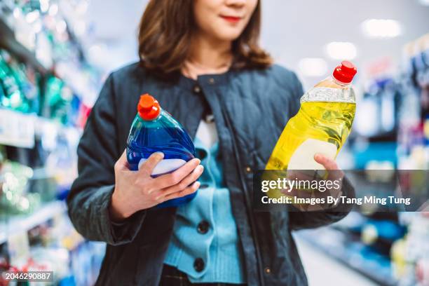 beautiful asian woman reading & comparing the product info of two bottles of detergent while doing groceries shopping in supermarket. consumer awareness concept. - cleaning equipment stock pictures, royalty-free photos & images