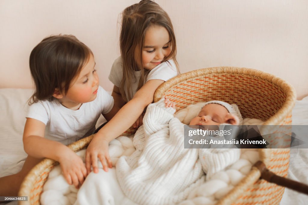 Two sisters playing and waching for newborn brother in whicker crib at home.