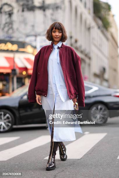 Flora Coquerel wears a burgundy oversized bomber jacket , a pastel pale blue long shirt / midi dress with a tie, a leather bag, knee high leather...