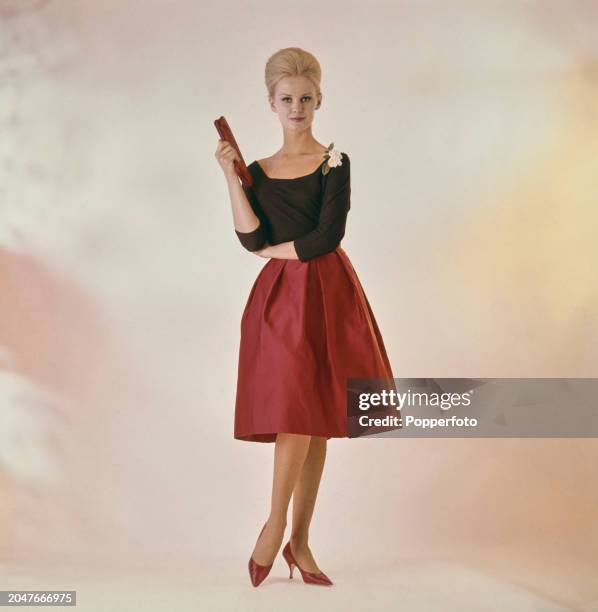 Posed studio portrait of a female fashion model wearing a scoop neck black top and red knee length box pleated skirt and matching red shoes, London,...