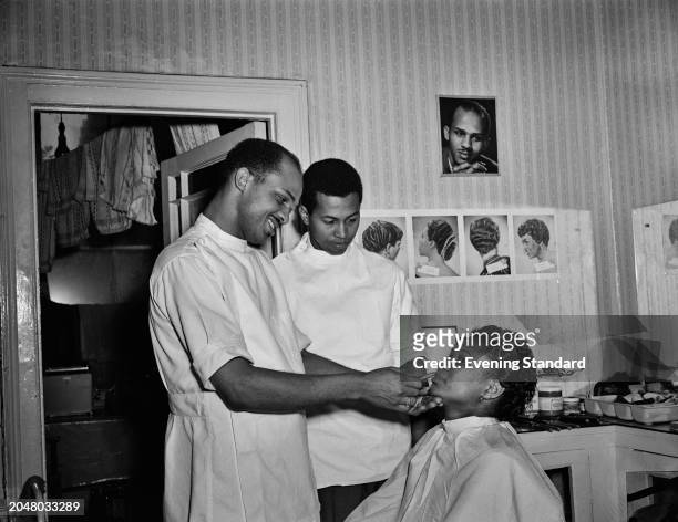 Jamaican hairdresser Roy Lando applies lipstick to a customer in his salon, August 22nd 1956. Behind him on a wall are photos of hairstyles.