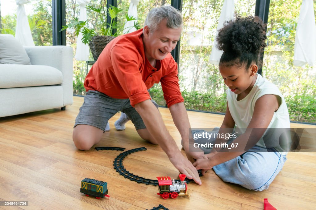Happy African American family, senior grandfather and his little black granddaughter enjoy playing toy train on floor while spending free time together at home.
