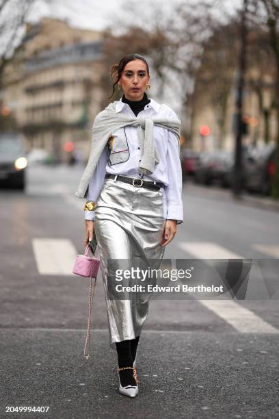 Gabriella Berdugo wears golden earrings, a white shirt, a gray wool pullover , a black leather belt, a silver midi shiny skirt , a golden bracelet,...