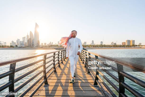 young man wearing keffiyeh and dishdasha and walking on the dock in kuwait city - kuwait tradition stock pictures, royalty-free photos & images