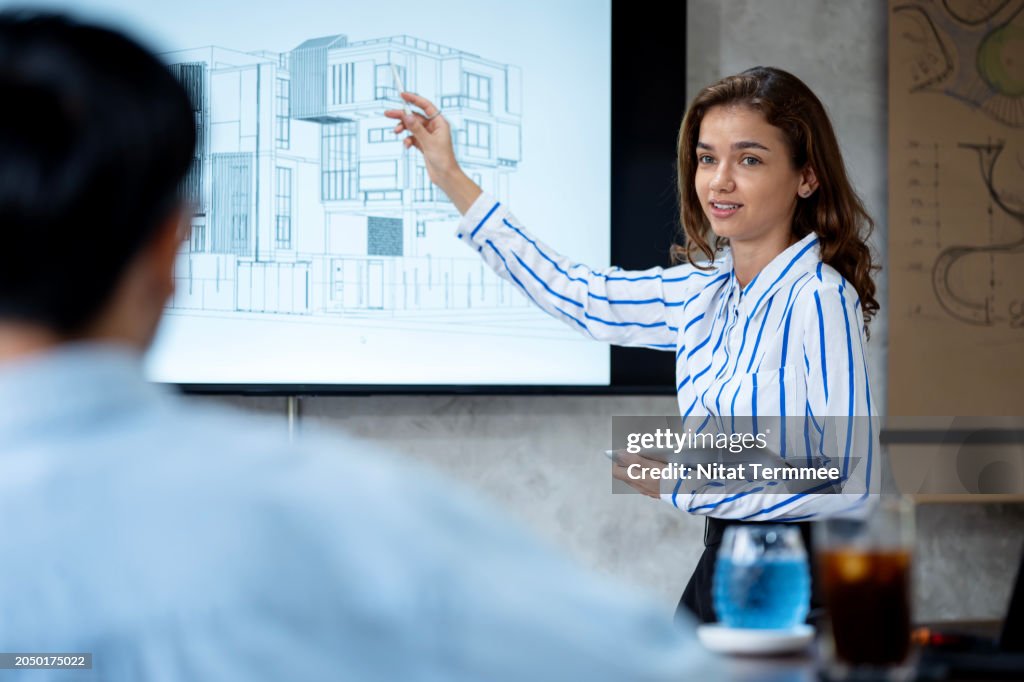 Managing Your Design Projects Process to Ensure Compliance. A female designer is discussing construction projects design with her team on a display LCD to ensure compliance in a modern office.