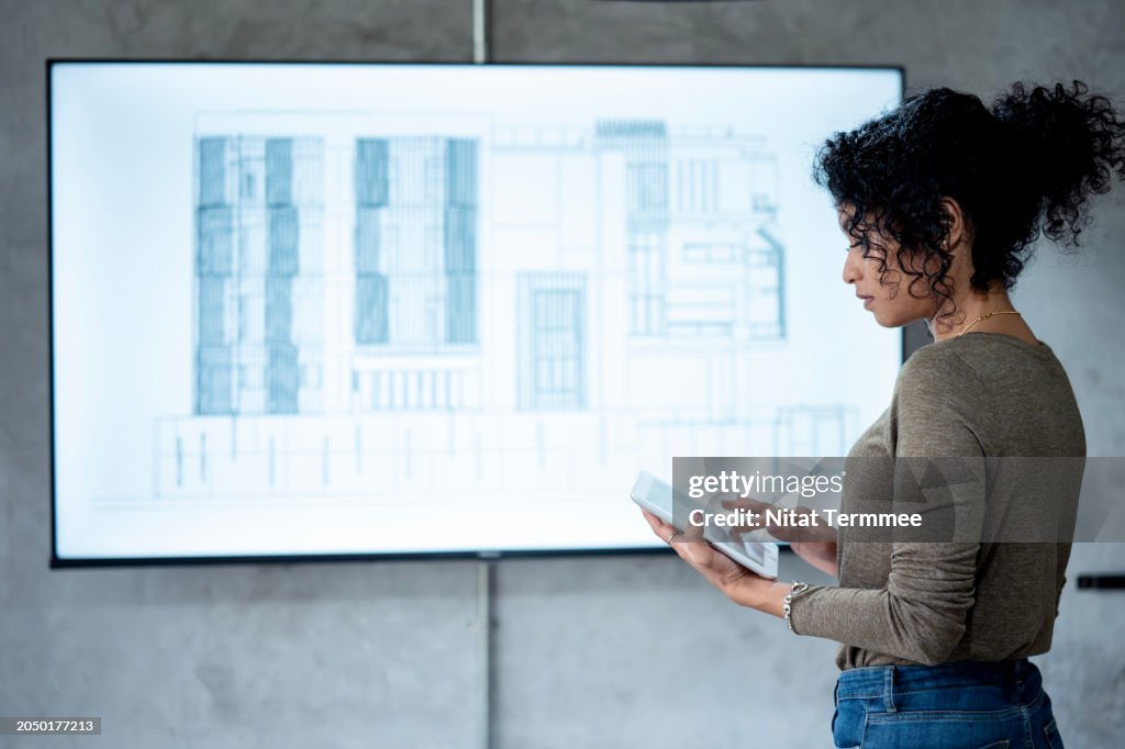 Energy-efficient Design and Construction to Towards a Sustainable Future. A female project leads a project design review in front of a digital blueprint on a device screen during the design phase in an architect's office.