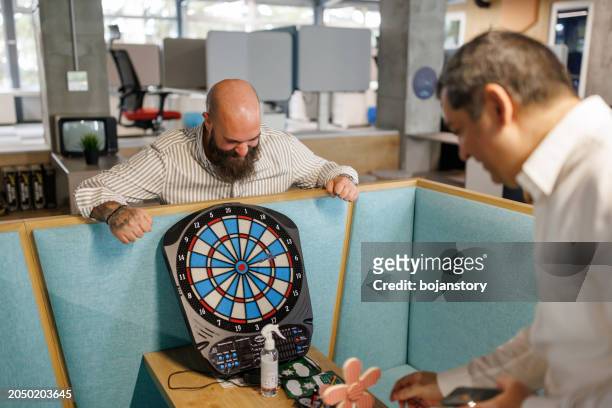 male business colleagues playing darts in modern office lobby - man darts stock pictures, royalty-free photos & images