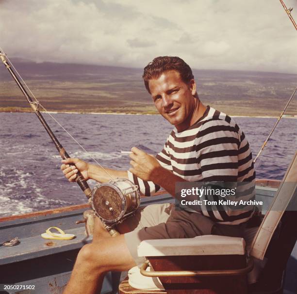 American football player and sports commentator Frank Gifford, wearing a black-and-white hooped top and grey shorts, smokes a cigarette as he...