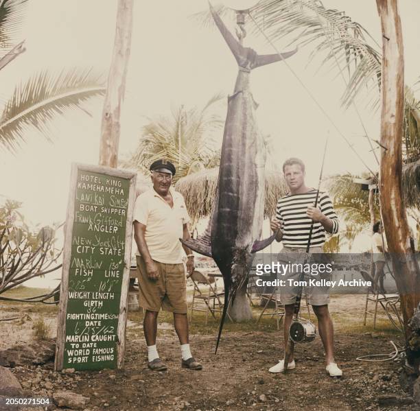 American football player and sports commentator Frank Gifford, wearing a black-and-white hooped top and grey shorts, poses among palm trees with a...