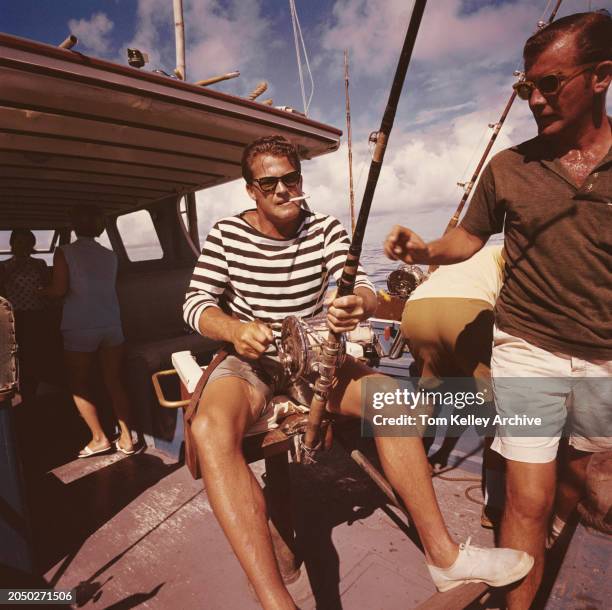 American football player and sports commentator Frank Gifford, wearing a black-and-white hooped top and grey shorts, smokes a cigarette as he...