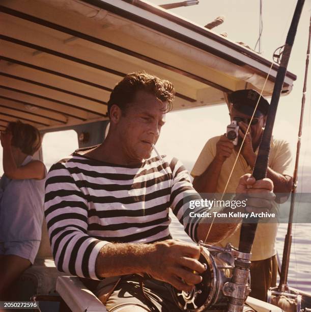 American football player and sports commentator Frank Gifford, wearing a black-and-white hooped top and grey shorts, smokes a cigarette as he...