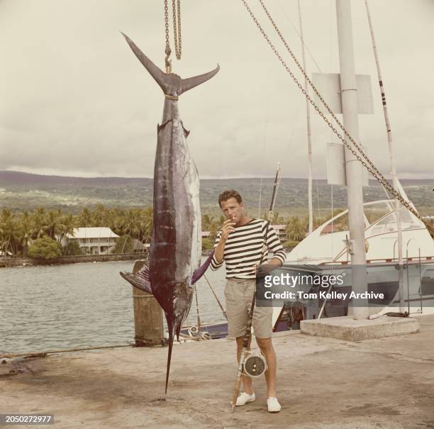American football player and sports commentator Frank Gifford, wearing a black-and-white hooped top and grey shorts, poses on the quayside beside a...