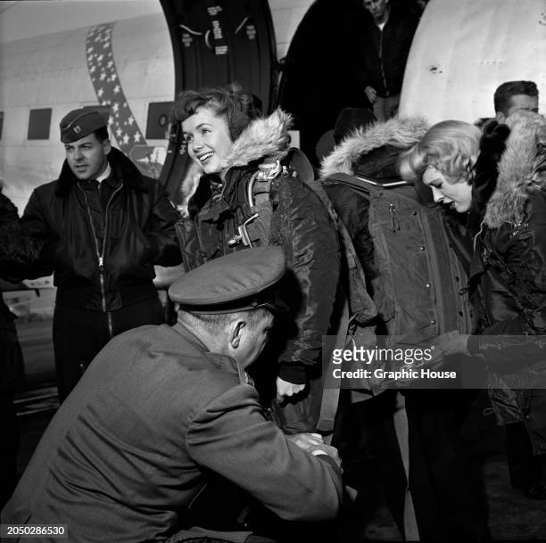 American actress and singer Debbie Reynolds, wearing a fur-trimmed flight jacket and a parachute, smiles as she is readied for her flight to attend a...
