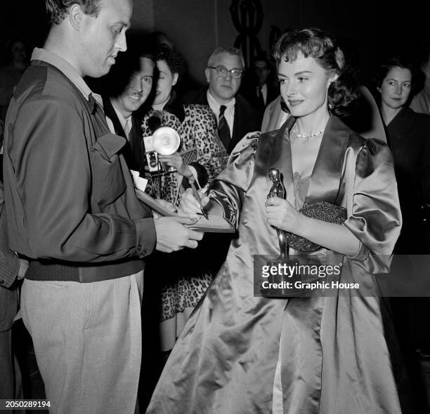 American actress Donna Reed, wearing a satin evening coat over her outfit, signs an autograph for a fan as she leaves the 26th Academy Awards, held...