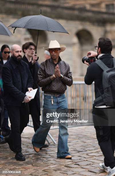 Pharrell Williams seen wearing beige large cowboy hat, black sunglasses with orange lenses, gold earrings and a gold necklace, Louis Vuitton brown...