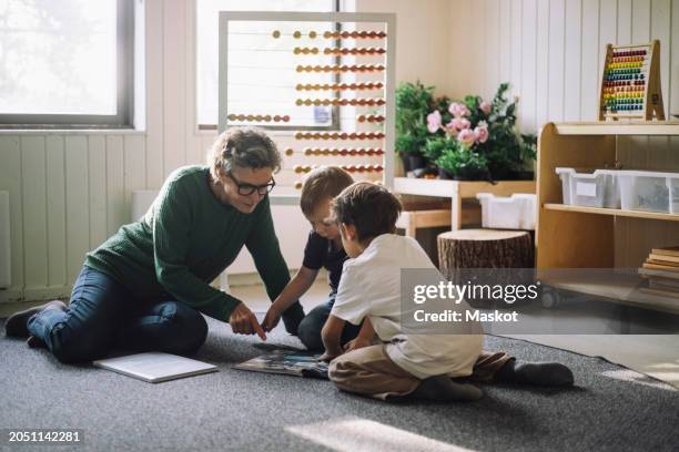 senior female teacher sitting with preschool children reading book at kindergarten - sitting on ground stock pictures, royalty-free photos & images